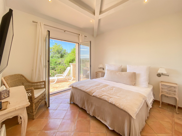 Bright and airy bedroom featuring a queen-size bed with beige linen, terracotta tiled flooring, white walls, and wooden ceiling beams, with French doors opening to a sunny balcony overlooking lush greenery. Experience tranquility in this sunlit bedroom, where natural light floods through large French doors that open onto a picturesque balcony, revealing a stunning view of verdant surroundings. The room is thoughtfully appointed with a comfortable queen-size bed draped in soft beige linens that invite relaxation. Terracotta tiles underfoot add warmth to the space, while the white painted walls and exposed wooden ceiling beams create a feeling of spaciousness and rustic charm. A wicker armchair and matching side tables enhance the room's cozy yet elegant ambiance, making it an ideal retreat for rejuvenation. This bedroom seamlessly blends indoor comfort with the beauty of nature, providing a peaceful escape from the everyday.