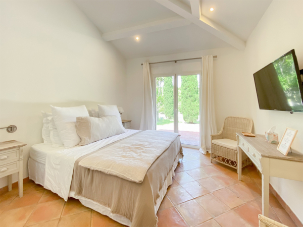 Spacious bedroom featuring terracotta floor tiles, a large bed with white and beige linens, a rattan chair, a wooden writing desk, and natural light streaming in through French doors opening to a garden. Discover the peaceful ambiance of this rustic bedroom designed for relaxation and comfort. The room is bathed in natural light that enhances the soft, neutral palette of whites and beiges. Terracotta tiles add warmth to the floor, complementing the quaint charm of the exposed white beams overhead. A cozy rattan chair sits beside a classic wooden desk, offering a perfect nook for reading or journaling. The plush bedding invites you to unwind, making it an ideal setting for a restful retreat.