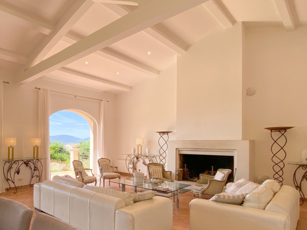 Luxurious living room in a Mediterranean-style villa featuring white sofas, a glass coffee table, wrought iron floor lamps, and a large arched window offering panoramic views of distant mountains. Step inside this elegant living room, designed with a sophisticated Mediterranean flair, captured in a villa that overlooks stunning mountain vistas. The room boasts high vaulted ceilings with white beams, enhancing the spacious feel, while soft natural light floods through a grand arched window, framing the breathtaking landscape beyond. Furnished with plush white leather sofas and a sleek glass coffee table, the space invites relaxation and comfort. Elegant wrought iron floor lamps add a touch of rustic charm. This serene setting is perfect for those who appreciate the fusion of luxury and natural beauty, making it an ideal inspiration for homeowners and interior designers aiming to create a tranquil, yet opulent living environment.