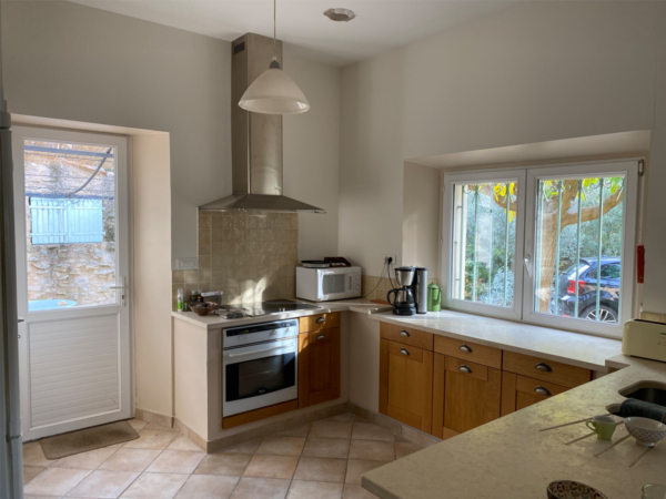 A well-lit kitchen space with wooden cabinets, modern appliances, and a view to the outdoors. This photograph captures a quaint and cozy kitchen area that beautifully melds rustic charm with modern functionality. Natural light streams through large windows, highlighting the warm wooden cabinetry and contrasting pleasantly with the sleek stainless steel appliances. The inviting space promises culinary adventures and warm family meals, with thoughtful touches like a hanging pendant light and stone wall adding character and depth. The view of a serene outdoor area suggests a harmony between the home's interior comfort and the beauty of the natural world just outside, inviting one to cook with inspiration drawn from both inside and out. This kitchen embodies a personal haven where every meal is both an act of creation and a moment to cherish.
