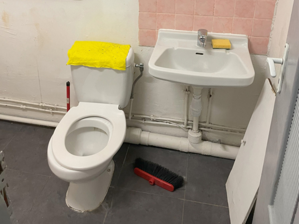 A basic bathroom setup with a white toilet and sink against a wall with exposed plumbing and patchy paint. This image presents a straightforward, no-frills bathroom with the essentials: a standard white toilet and a pedestal sink. The exposed plumbing and partially painted walls suggest a work in progress or a space maintained for sheer functionality over aesthetic appeal. A bright yellow cloth on the toilet seat and a sponge on the sink indicate regular use and cleaning. This bathroom embodies practical simplicity and stands as a canvas for future renovation, ready to be transformed into a more inviting and modern facility.
