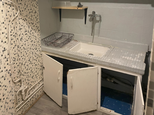 An old laundry area featuring a large sink with a mosaic backsplash, flanked by worn cabinetry under a tiled countertop. This image captures the essence of a bygone era in a laundry space that bears the marks of many uses. The large basin sink and its mosaic tile backdrop speak of functionality and a no-nonsense approach to household chores. The cabinet doors hang open, revealing the need for attention and care. With a hint of wear and potential water damage, it's a space ripe for restoration, waiting to be infused with new life and modern functionality while preserving its rustic charm. It's a testament to the resilience of well-used spaces and the endless possibilities that lie in refurbishment.