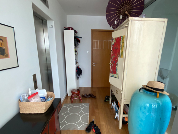 A residential entryway filled with everyday items including a coat stand, an umbrella, a storage cabinet, and a mirror. This image captures a lived-in entryway, a testament to the comings and goings of daily life. Directly ahead, a wooden door signals passage to the home's private sanctuaries. To the right, a cream-colored cabinet adorned with an intricate red cloth stands beside a bold turquoise vase, the latter adding a dash of color to the space. An assortment of personal items, such as hats and bags, are casually draped over a standing coat rack, evoking a sense of arrival or departure. A traditional umbrella opens up towards the ceiling, perhaps left out to dry, adding an element of charm to the corner. The scene is grounded by a practical grey rug, its geometric pattern offering a subtle decorative touch. The atmosphere is one of welcoming disorder, a space where functionality and personal touches blend to create a snapshot of home life.