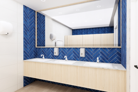A stylish female washroom interior showcasing a large mirror, white sinks with modern fixtures, and vibrant blue herringbone tiles contrasted with light wooden cabinetry. This female washroom interior combines elegance and modern design. The eye is immediately drawn to the vibrant blue herringbone tiles that adorn the wall, providing a dynamic backdrop to the sleek white sinks. Above, a large, frameless mirror spans the length of the vanity, reflecting the well-lit space and amplifying its airy feel. Modern fixtures in clean lines are mounted onto the wall, maintaining the contemporary aesthetic. Below, the light wooden cabinetry brings warmth to the room, softening the boldness of the tiles and complementing the neutral tones of the flooring. This design is not only visually pleasing but also serves to create an inviting and comfortable space for its users.