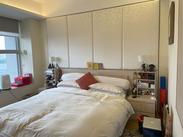 A well-lived bedroom with a king-size bed covered in white linens, with a patterned wallpaper headboard. Side tables are filled with personal belongings and lamps. A single framed picture hangs on the wall, and various everyday items are scattered around, giving a personalized touch to the space. This bedroom presents a lived-in comfort with its large bed dressed in inviting white linens. The headboard, simple in design, is flanked by two practical bedside tables cluttered with everyday items and personal knick-knacks, depicting a scene of daily life. The wall behind the bed features a subtle patterned wallpaper that brings a touch of elegance without overpowering the room’s casual atmosphere. Each side table hosts a lamp, providing ample light for nighttime reading or relaxation. Various personal items and colorful accents reflect the room's individual character and usage, while a singular framed piece of art on the wall adds a cultured touch to the otherwise utilitarian space.