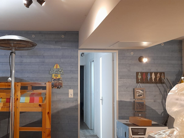 A room with a wooden dining set against a wall with grey faux wooden paneling and surf-themed decor. This dining area showcases a relaxed urban surf theme, evidenced by the grey faux wood paneling that sets a laid-back tone. A robust wooden bunk bed with bright multi-colored bedding adds a playful touch, indicative of a casual beach hostel vibe. To the right, the surf theme is reinforced by a vibrant "Surf Shack" wall sign and a decorative piece featuring a lineup of colorful surfboard fins. The room is illuminated by a modern heater lamp and recessed ceiling lights, balancing practicality with an inviting ambiance. A wooden cabinet with a display of neatly organized beach-inspired decor items, along with a simple yet functional white door, provide practical elements without sacrificing style. The overall look suggests a space perfect for young, adventure-seeking spirits with a penchant for the surfer lifestyle and urban design.