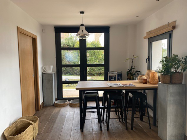 A cozy dining area with a rustic wooden table and black chairs, large windows with garden views, and potted plants adding a touch of greenery. This inviting dining space exudes a rustic charm, featuring a sturdy wooden table surrounded by sleek black chairs that provide a modern contrast. Ample natural light floods in through large glass windows with black frames, offering a view of the greenery outside, enhancing the room's connection with nature. An assortment of potted plants on a concrete-like console brings life and freshness to the area. Strategically placed woven baskets on the warm wooden floor and a simple pendant light hanging from the ceiling contribute to the room's homey and welcoming atmosphere. This dining nook is a perfect blend of simplicity, warmth, and functionality, making it an ideal spot for leisurely meals and conversations.