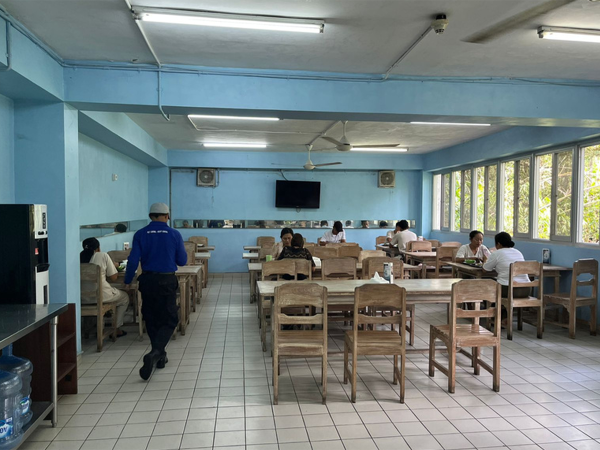 Spacious cafeteria with light blue walls, wooden tables and chairs, and people dining, illuminated by natural light from large windows. Embark on a visual tour of this casual dining hall, where the simple charm of communal eating is celebrated. The room is bathed in a soothing light blue hue, creating a calm and welcoming atmosphere. Sturdy wooden tables and chairs are neatly arranged, inviting diners to enjoy their meals in good company. Patrons, in varying attire from work uniforms to casual clothes, are captured in moments of relaxation and engagement, with some focused on their food and others deep in conversation. The hall's fluorescent lighting fixtures ensure a well-lit environment, complemented by an abundance of natural light streaming through the generously sized windows that offer a view of the greenery outside. A water cooler and a refrigerator stand at the ready, catering to the diners' needs. This space, with its unpretentious setup, reflects the everyday interactions and the shared experiences that form the heart of a communal dining area.