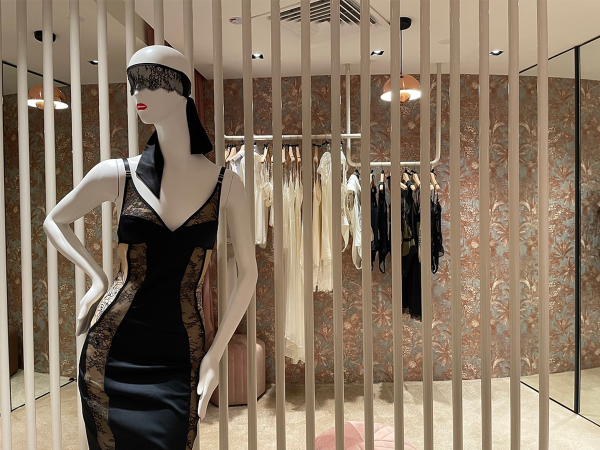 Mannequin in a boutique dressed in a black lace-trimmed dress, posing elegantly behind vertical bars, with a backdrop of floral wallpaper and delicate garments. Captured in this image is the essence of high-fashion display — a stunning mannequin dressed in a black dress with intricate lace trim, making a bold statement behind a chic set of vertical bars. This striking pose, reminiscent of haute couture allure, is set against a backdrop of opulent floral wallpaper that adds a romantic touch to the boutique's ambiance. Beyond the bars, the room holds a soft glow from the warm lighting that spotlights an array of delicate garments in creams and blacks, hanging gracefully and promising an exclusive selection for the discerning shopper. The mannequin's pose, both dynamic and statuesque, invites onlookers to admire the fusion of contemporary fashion and classic elegance. Each detail in this boutique's display, from the lace blindfold on the mannequin to the curated fashion pieces, is carefully designed to capture the imagination and evoke a sense of luxury and refined taste.