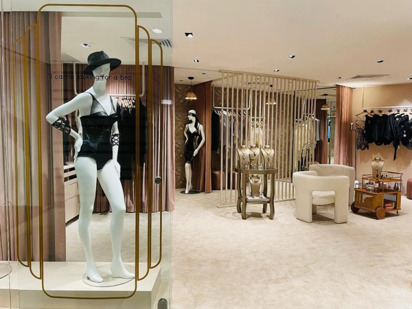 Fashionable mannequin posed in a boutique window, styled in a corset and hat, against the soft ambiance of the store's interior featuring gold accents and lush drapery. Stepping into the frame with undeniable presence, a mannequin in this boutique window exudes modern chic, dressed in a striking corset, accentuated by a poised black hat that adds a touch of enigmatic flair. The store's interior sets a luxurious stage with its soft, plush carpeting and a harmonious blend of golden accents that grace the space — from the sleek vertical bars to the elegant fixtures. Draped curtains in a gentle blush tone offer a contrast to the bold fashion statement, encapsulating the boutique's commitment to providing a sophisticated shopping experience. In the background, a refined selection of garments awaits the discerning eye, while a round table adorned with jewelry and a vintage-inspired display cart add character to the boutique. The ambiance is one of cultivated taste and style, promising an intimate and personalized encounter with fashion for every visitor.