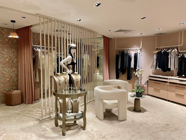 Chic boutique interior with an elegantly posed mannequin behind vertical golden bars, surrounded by high-end clothing racks and plush furniture. Welcome to an epitome of sophistication within this boutique's luxurious interior. Dominating the scene is an elegantly posed mannequin, styled with exquisite lingerie and accessories, strategically placed behind vertical golden bars that evoke exclusivity and allure. The backdrop is a rich tapestry of floral wallpaper, adding depth and texture to the setting. To the right, the interior opens up to showcase meticulously arranged high-end clothing racks, with garments in varying shades of black and beige, promising a treasure trove of fashionable finds. In the foreground, a circular glass table presents a selection of fine jewelry, anchoring the space as a focal point of refined taste. The seating arrangement features a modern, sculptural chair and a subtle, yet luxurious, ottoman, inviting a moment of relaxation in the plush ambience. Soft carpeting underfoot and strategically placed lighting ensure that each product shines, while a nearby potted orchid adds a breath of life and elegance to the composition. This boutique’s design seamlessly marries fashion with art, creating an environment that is as inviting as it is impressive, waiting to enchant those who appreciate the finer things in life.