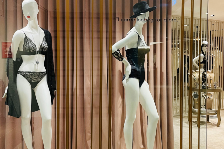 Two mannequins in a boutique window display, one dressed in a black lace bodysuit and sheer robe, the other in a corset with a black hat, against a backdrop of elegant brown curtains. Enter a realm of high-end fashion with this boutique window display, showcasing the latest in lingerie elegance. On the left, a poised mannequin stands adorned in a luxurious black lace bodysuit complemented by a flowing sheer robe, epitomizing the blend of comfort and sophistication. Beside it, a mannequin strikes a confident pose, its silhouette accentuated by a sleek, figure-hugging corset, topped with a stylish black hat that adds an aura of mystery. The display is framed by sumptuous brown curtains with soft, vertical pleats that provide a textured backdrop, while the quote "I came looking for a bra" playfully adorns the glass, inviting passersby to explore the allure within. Golden arches crown the display, adding a touch of opulence and drawing the eye to the craftsmanship of the lingerie pieces, making it a captivating sight for fashion enthusiasts and casual onlookers alike.