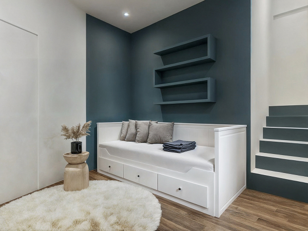 A minimalist bedroom with a white daybed, a blue accent wall, a white furry rug, and wooden shelves.