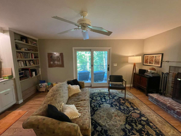 Living room before redesign, showing a patterned sofa, bookshelf filled with books, a record player, a traditional fireplace, and a door leading to an outdoor deck.